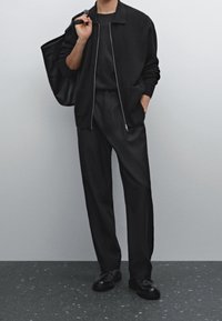 Schwarze Strickjacke mit Reißverschluss, schwarzes Hemd und maßgeschneiderte schwarze Hose. Accessoires sind eine strukturierte schwarze Tragetasche und Loafers. Weicher, strukturierter Look.