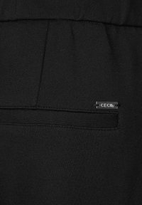 Zwarte joggers van stof met een gladde textuur, voorzien van een achterzak en een klein metalen label met de tekst "CECIL." Het ontwerp is eenvoudig en gestroomlijnd.