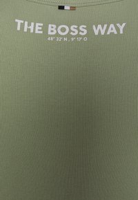 Zielony top z żeberkowanej tkaniny z białym napisem „THE BOSS WAY” oraz współrzędnymi geograficznymi, z małą, wielokolorową metką przy dekolcie.