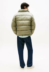 Chaqueta acolchada de color verde oliva con costuras horizontales, cuello alto y bolsillos laterales, combinada con pantalones vaqueros de denim azul oscuro y zapatillas blancas.
