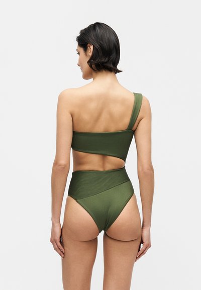 Γυναίκα που φοράει ένα πράσινο, ριπ, μπικίνι τοπ με έναν ώμο και μαγιό σλιπ με ψηλή μέση, γυρισμένη προς τα πίσω σε ένα απλό ανοιχτό φόντο.