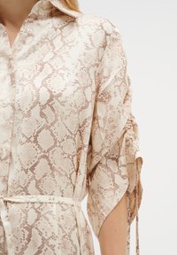 Beige paita kuviolla, jossa on nappikaulus, pehmeä kangas, kolme neljännestä hihoista ja nauhavyö korostamaan vyötäröä.