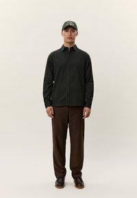 Άνδρας που φοράει πουκάμισο με κατακόρυφες ρίγες σε σκούρο πράσινο, καφέ παντελόνι, σκούρα παπούτσια και πράσινο καπέλο μπέιζμπολ, στέκεται μπροστά σε απλό φόντο.