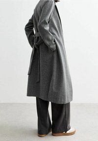 Abrigo gris de espiga con cinturón atado, mangas largas y ajuste relajado, combinado con unos pantalones de pierna ancha gris oscuro y calzado de color claro.