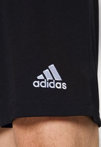 Pantaloni corti neri Adidas con logo ricamato bianco ben visibile sopra il ginocchio di una persona.