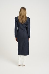 Pitkä tummansininen denim-mekko, rakenteelliset hartiat, slim fit -malli, jossa on takasauman yksityiskohtia. Mallilla on päällään valkoiset korkeat naskalit.