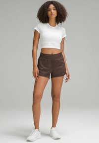 Barna sportos rövidnadrág elasztikus derékrésszel, egy világos fehér crop top-pal párosítva. A modellen fehér sportcipő van. Simas textúrájú anyag.