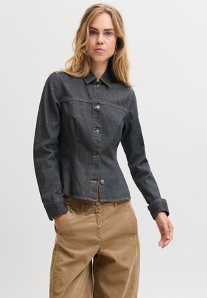 Une femme porte une veste en jean foncé boutonnée et un pantalon beige devant un fond clair et uni.