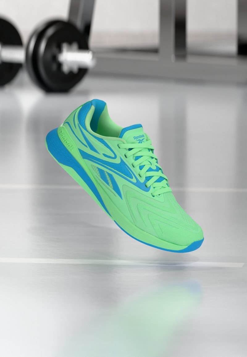 Φωτεινό πράσινο και μπλε αθλητικό παπούτσι Reebok που αιωρείται πάνω από μια αντανακλαστική γυμναστήρια επιφάνεια με θολές αλτήρες στο φόντο.