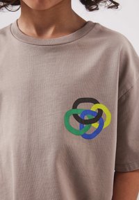 Personne portant un t-shirt taupe avec quatre anneaux entrelacés en noir, vert, bleu et jaune imprimés sur la poitrine.