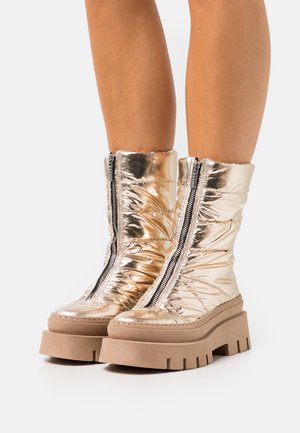 Metalen gouden enkelboots met een gequiltde textuur, contrasterende zwarte ritsen en dikke beige platformzolen. Over het algemeen een modern ontwerp.