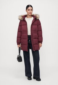 Kvinde iført en bordeauxfarvet polstret jakke med pelskrave, hvid sweater, mørk bred benjeans, sorte plateau-sko og holder en lille sort håndtaske.