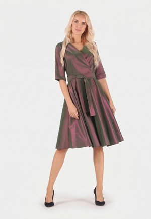 Frau mit langen blonden Haaren, die ein glänzendes, knieumspielendes Wickelkleid in Lila-Grün mit Gürtel und schwarzen High Heels trägt, vor weißem Hintergrund stehend.