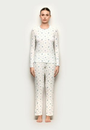 Mujer descalza que lleva puesta una blusa de manga larga blanca y pantalones con pequeños estampados florales azules y morados sobre un fondo liso y claro.