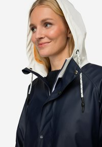 Námořnický voděodolný pláštěnka s bílou nastavitelnou kapucí, cvokovými knoflíky a kontrastními vzorovanými šňůrkami, s hladkou, lesklou strukturou.