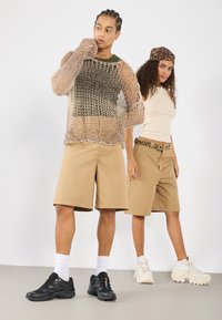 Un short beige, un débardeur vert et un pull en maille transparente mettent en valeur le modèle masculin, tandis que la femme porte un haut crème et une ceinture en imprimé léopard.