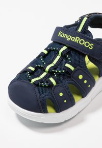 Navy blue sandal with mesh material, green accents, adjustable strap, and reinforced toe cap. Features a white rubber sole and perforated sides.