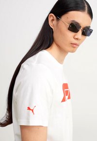 White cotton t-shirt with red Puma logo on the front and red Puma cat logo on the left sleeve; model wearing angular black sunglasses.