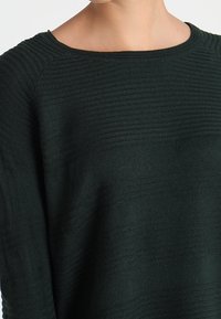 Mörkgrön stickad tröja med en strukturerad ribbad design, rund halsringning och avslappnad passform. Har vida ärmmuddar och subtila diagonala linjer.