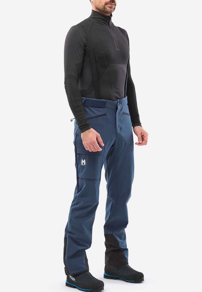 Couche de base noire à manches longues avec col zippé, associée à un pantalon imperméable bleu avec plusieurs poches et des accents noirs. Porte des bottes noires.