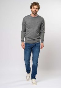 Mies kävelee eteenpäin harmaa neule päällä, siniset farkut ja valkoiset tennarit, yksinkertaista vaaleaa taustaa vasten.