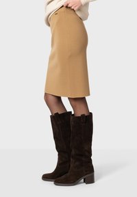 Jupe camel jusqu'aux genoux en tissu texturé, associée à des bottes en suède marron foncé hautes avec des talons blocs et des accents de tirette.