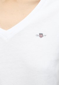 T-shirt blanc à col en V avec un petit logo bouclier brodé comprenant des étoiles et des rayures, placé sur la partie gauche de la poitrine.