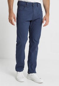 Pantalon en coton bleu marine à coupe droite, doté de poches avant, d'une fermeture boutonnée, porté avec des baskets blanches. Texture lisse.