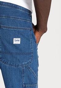Pantalones de trabajo de mezclilla en azul medio que presentan un logo en parche, múltiples bolsillos, costuras contrastantes y un diseño de ajuste relajado.