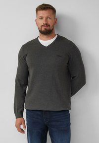 Grijze V-hals trui van zacht materiaal, met lange mouwen en een subtiele logo op de borst, gecombineerd met donkere jeans.