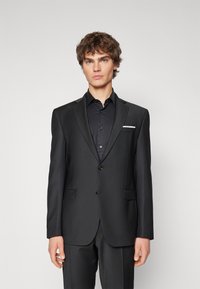 Czarna marynarka w eleganckim kroju, z klapami w kształcie notcha, na dwa guziki i białą poszetką. Materiał ma gładką, matową fakturę.