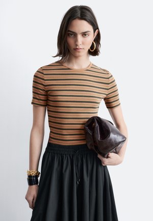 Jonge vrouw met kort donker haar, gekleed in een bruin-zwart gestreept shirt en een zwarte rok, terwijl ze een slungelige donkerbruine leren tas vasthoudt.