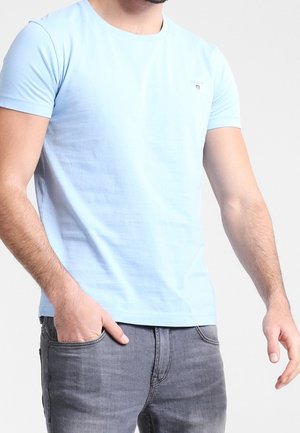 Homme portant un t-shirt bleu clair à manches courtes avec un petit logo sur la poitrine et un jean gris foncé, une main dans la poche.