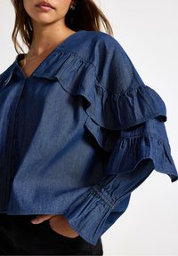 Denimskjorte med V-hals, volanter ved ærmerne og knapdetaljer i en dyb blå farve. Løs pasform med elastiske manchetter.