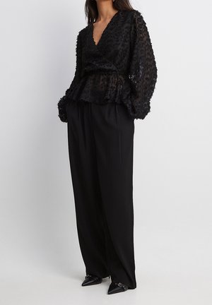 Femme portant un chemisier cache-cœur texturé noir à manches longues bouffantes, un pantalon large noir ample et des chaussures noires pointues avec des boucles.