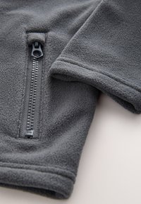 Gros plan sur la manche d'une veste polaire grise avec une poche zippée verticale, montrant un tissu doux et texturé ainsi que des coutures soignées.