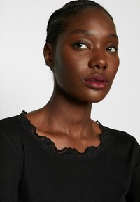 Top nero a coste con scollo in pizzo. Presenta un bordo ondulato e una consistenza morbida, design aderente che esalta la zona del colletto.