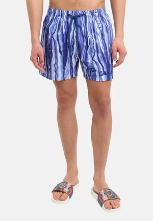 Homme portant un short de bain bleu et blanc à motifs avec un cordon et des sandales slides colorées sur un fond uni.