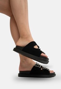 Sorte slide-on sandaler lavet af suede, med udskæringer, to justerbare spænder og en tykk gummisål for bedre traction.
