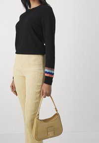 Kobieta w czarnym, długim rękawie z wielokolorowymi paskami na mankietach oraz beżowych, teksturowanych spodniach, trzymająca małą, kawową torebkę ze wzgórzem krokodyla.