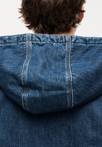 Φούτερ με κουκούλα από τζιν ύφασμα, με κορδόνια στην κουκούλα, μπλε ύφασμα, εμφανείς ραφές και χαλαρή εφαρμογή, φωτογραφημένο από πίσω.