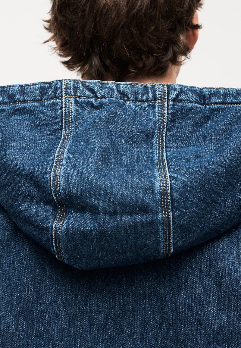 Denim hættetrøje med snøre i hætten, med blå stof, synlig syning og en afslappet pasform, set bagfra.
