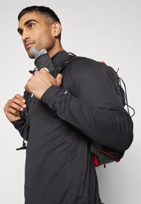 Zwarte sporttop met lange mouwen, textuurstof en een ritskraag. De persoon houdt een grijze waterfles vast die is bevestigd aan een zwart en rood rugzak.