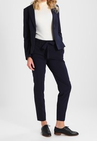 Marineblauw pak met krijtstreep, bestaande uit een getailleerd colbert en bijpassende taps toelopende broek, gecombineerd met een crèmekleurige geribbelde coltrui en zwarte nette schoenen.