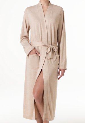 Accappatoio beige a maniche lunghe con cintura annodata a fiocco, due tasche frontali e spacco laterale che lascia intravedere una gamba piegata.