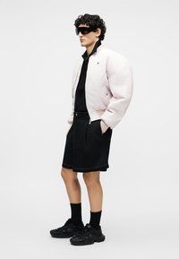 Άνδρας στέκεται φορώντας υπερμεγέθη ανοιχτό ροζ μπουφάν, μαύρο πουκάμισο, μαύρα σορτς, μαύρες κάλτσες, μαύρα αθλητικά παπούτσια και μεγάλα μαύρα γυαλιά ηλίου.