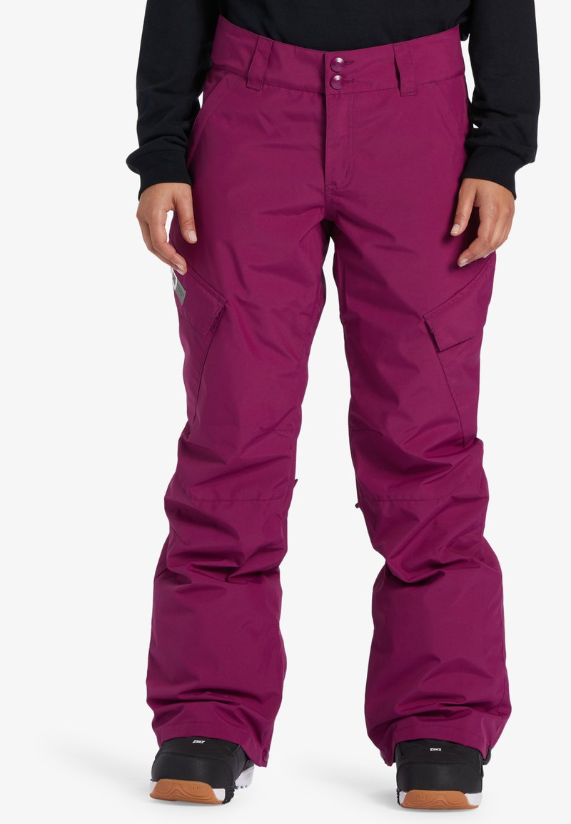 Fuchsia sneeuwbroeken gemaakt van waterdicht materiaal, voorzien van een verstelbare tailleband, ritszakken en een losse pasvorm. Zwarte schoenen eronder.
