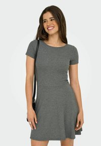 Jeune femme souriante, portant une robe grise à manches courtes en côte, des boucles d'oreilles créoles et portant un sac à épaule noir.