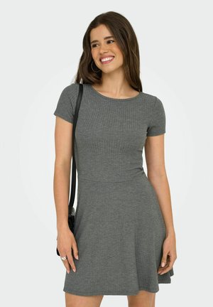 Jeune femme souriante, portant une robe grise à manches courtes en côte, des boucles d'oreilles créoles et portant un sac à épaule noir.