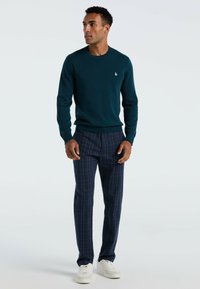 Pull en maille bleu canard, col rond, coupe slim. Pantalon à carreaux navy, coupe droite, texture subtile. Baskets blanches à profil bas.